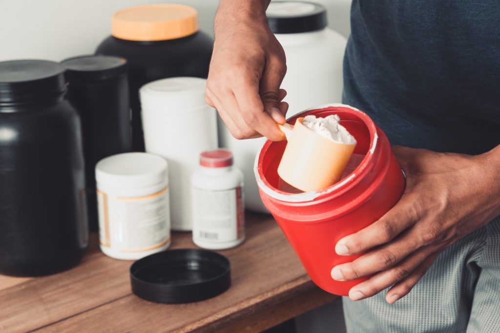 Hand scooping creatine powder from a jar with a measuring scoop, dietary supplements in the background